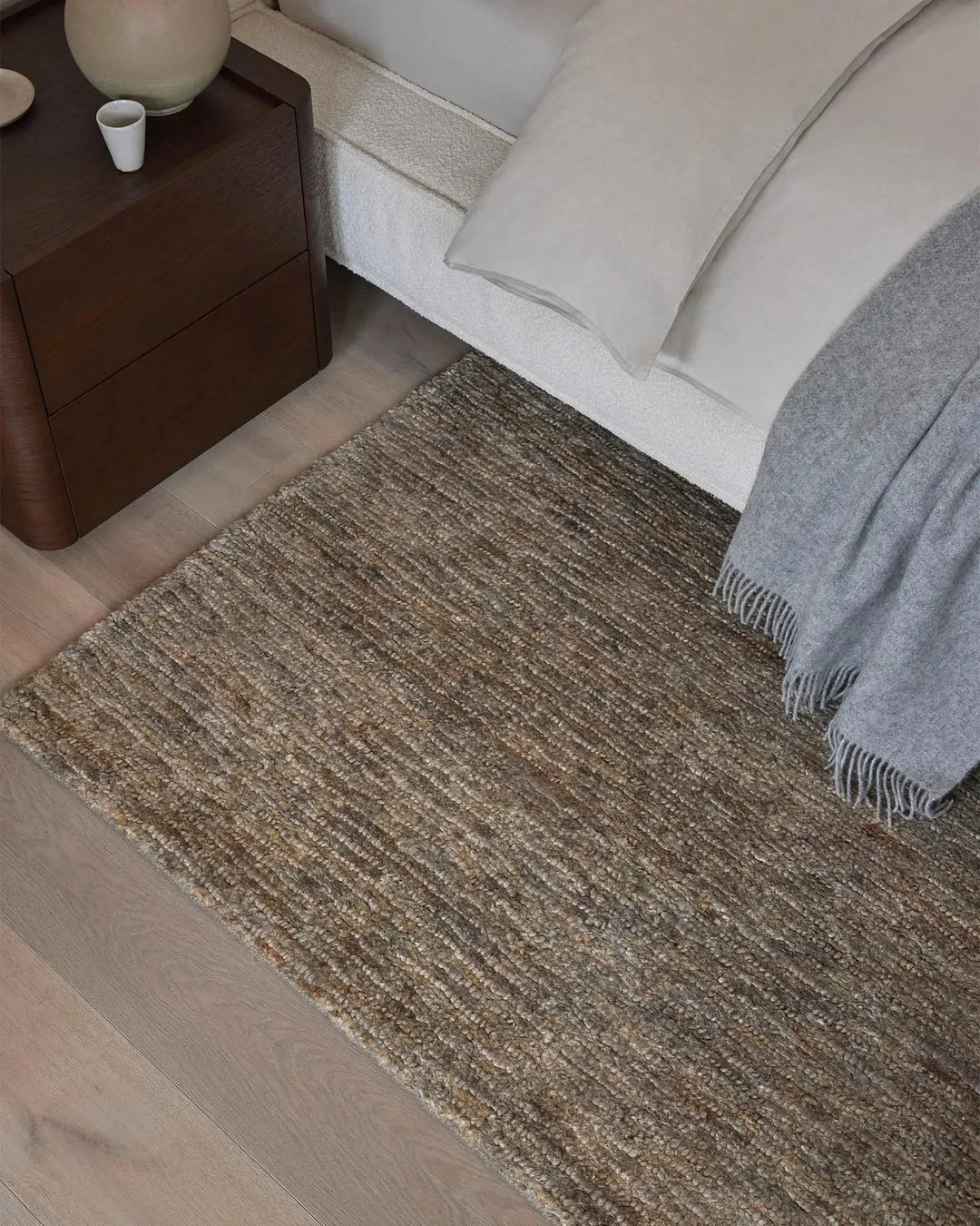 Brown textured jute rug on a wooden floor with a bed and nightstand in the background.