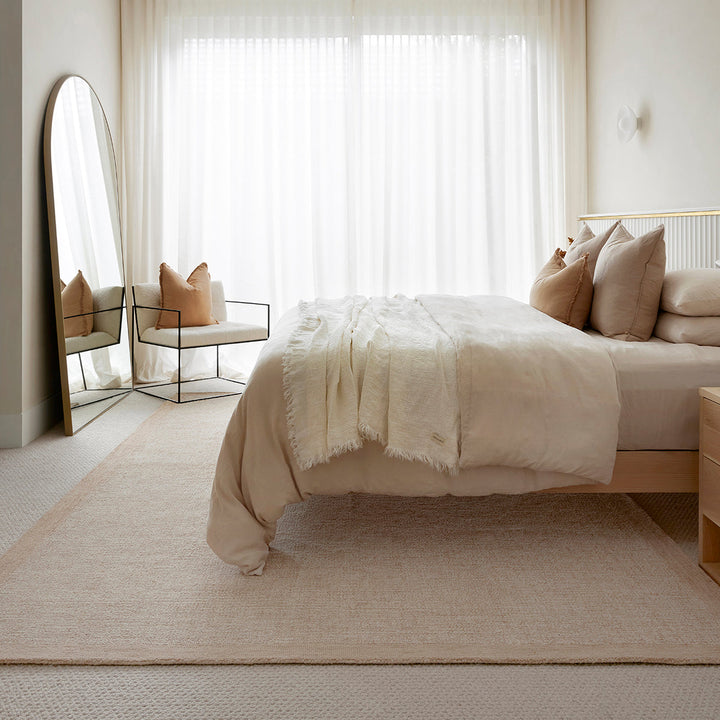 A styled bedroom scene featuring a light beige woven area rug under a bed with neutral bedding, a matching armchair beside a full-length floor mirror, and soft white sheer curtains in the background.