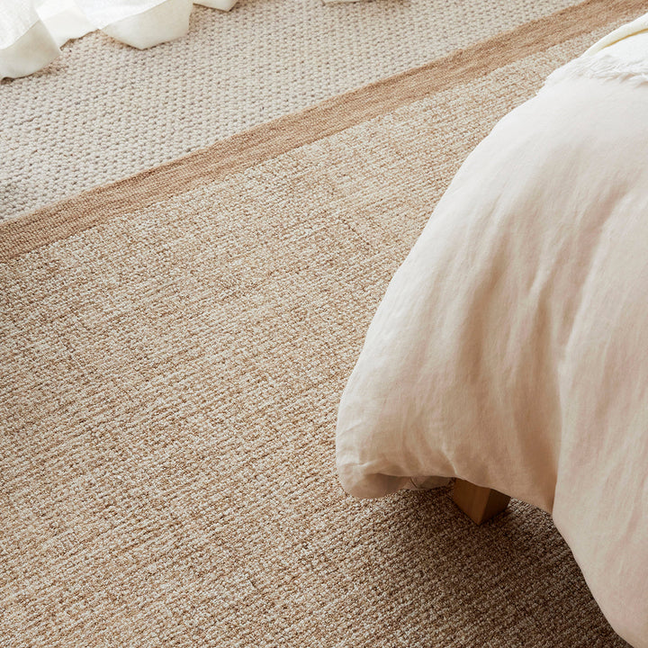 A close-up of a woven beige area rug with a textured basketweave pattern and a contrasting natural jute border, styled in a bedroom setting next to a bed with white bedding.