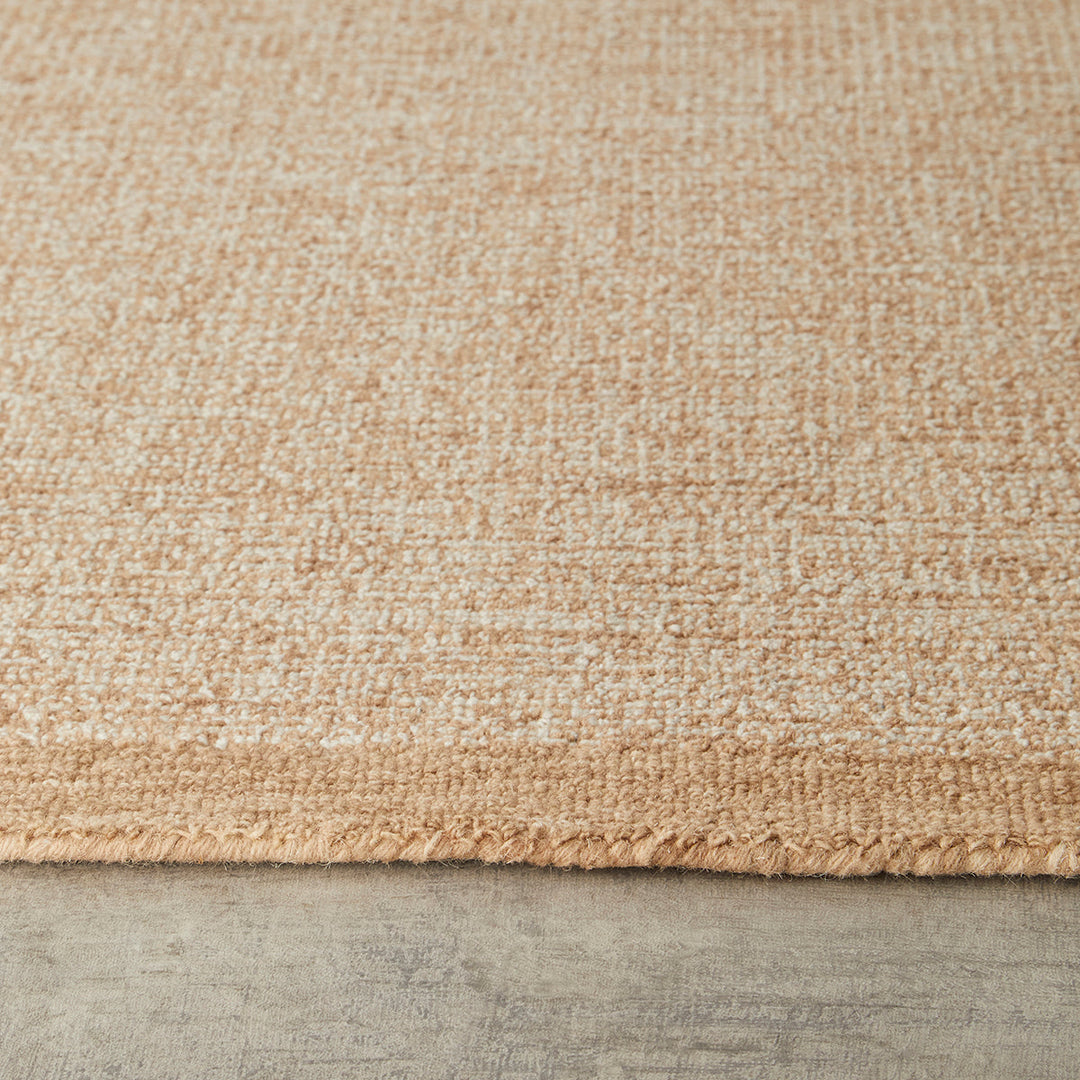 Close-up of a woven rug or textile in a warm beige/tan color with a short fringe along one edge, shown against a gray floor surface.