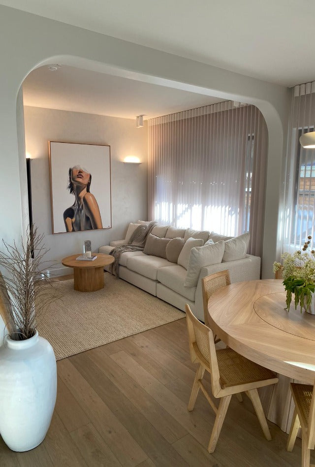 A well-lit living room featuring a beige wool rug under a coffee table with a framed artwork above the sofa, to the left a large white vase, and wooden dining chairs at the table.