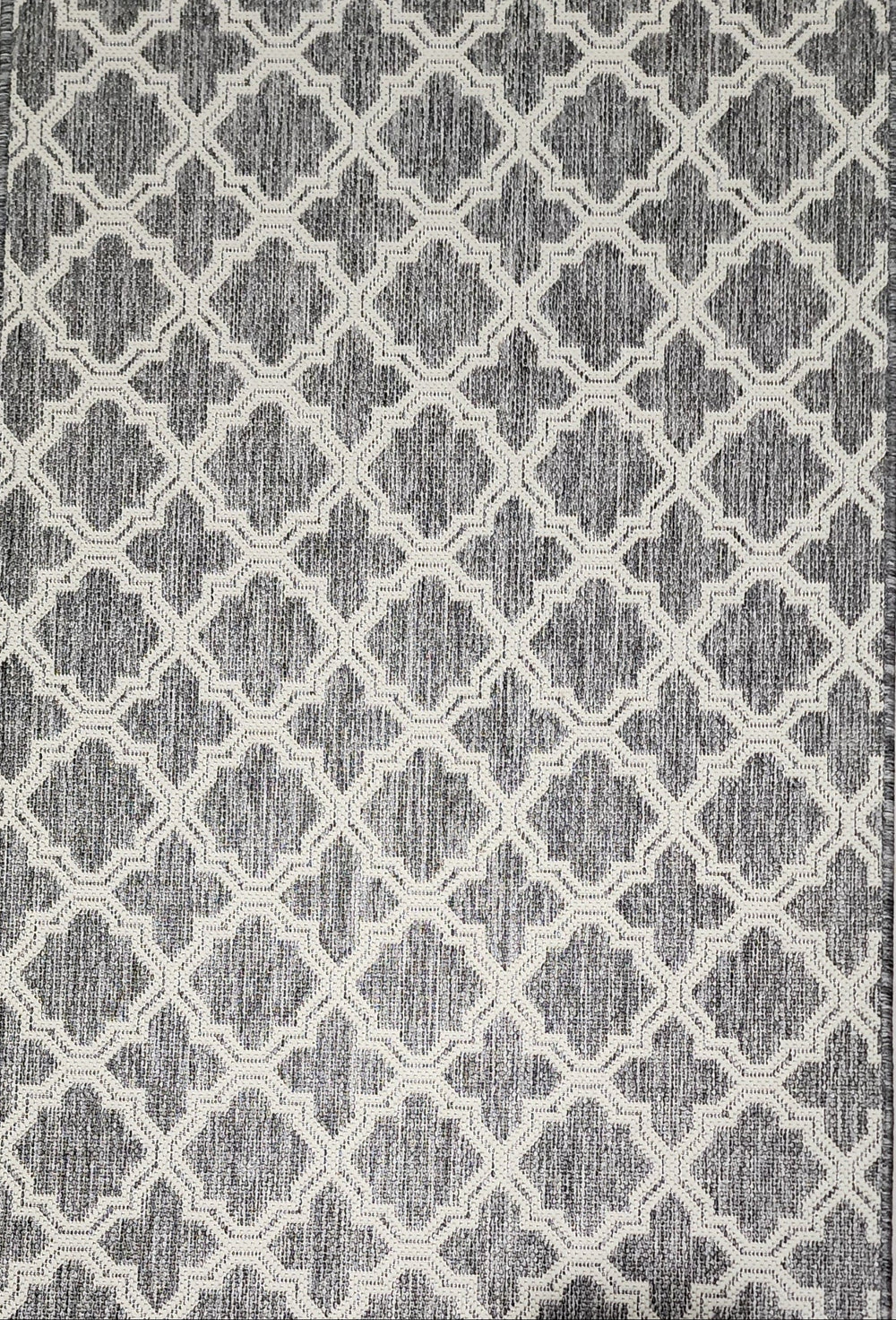 A rectangular area rug with a grey and off-white geometric trellis pattern, shown from a top down perspective on a light tiled floor with the feet of a person visible at the bottom edge.