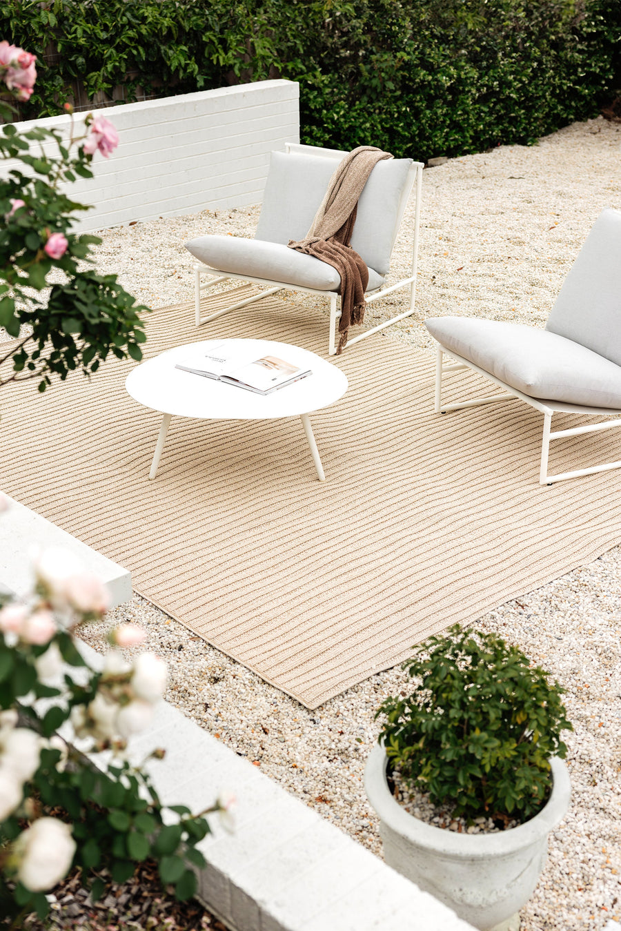 A taupe indoor/outdoor rug with a textured linear design, displayed in a garden setting with outdoor furniture set on top of it.