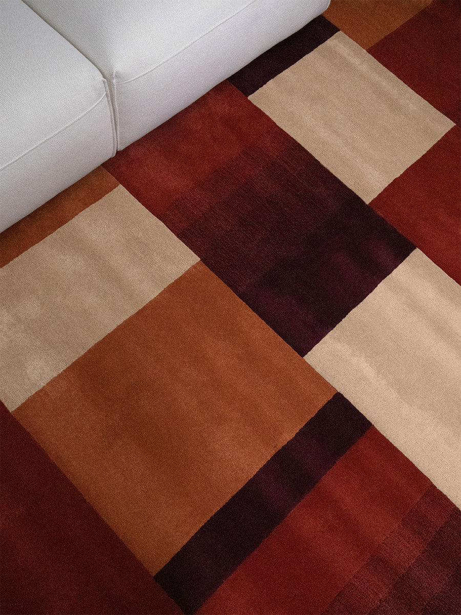 A close-up of a geometric area rug with a patchwork design in rust red, deep burgundy, tan, and cream, shown on a floor next to a white sofa.
