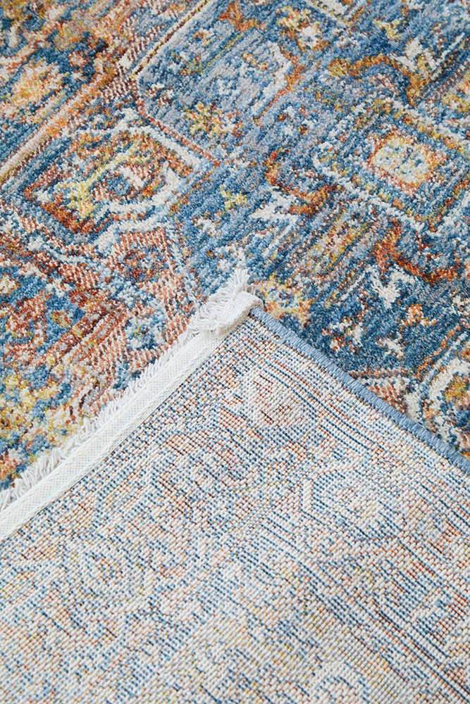 Close-up of a patterned area rug showing a blue, beige, and rust ornamental design with white fringe on one side.