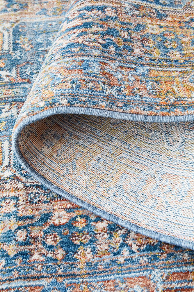 A close-up of a folded area rug showing its detailed traditional pattern in shades of blue, beige, and rust orange, with the underside and fringed edge visible.