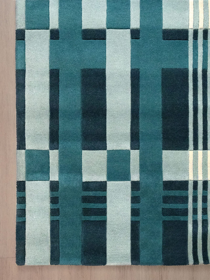 A close-up of a geometric area rug with a modern plaid pattern in shades of teal, navy, and light blue, laid on a light wood floor.