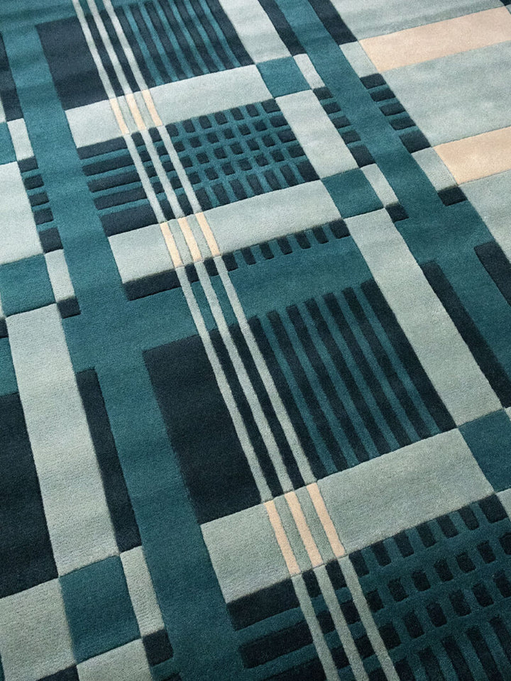 A close-up of a geometric area rug featuring a modern plaid pattern in shades of teal, slate blue, charcoal gray, and light beige.
