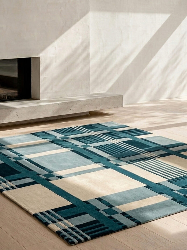 A modern area rug with a geometric plaid pattern in teal, slate blue, cream, and gray, styled on a light wood floor in a minimalist room with a concrete fireplace and warm natural light.