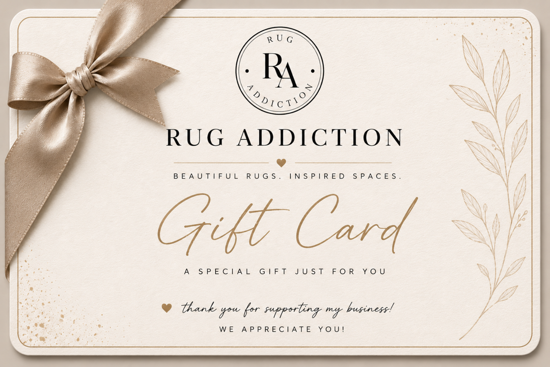 A square gift card from Rug Addiction with a beige satin ribbon bow in the top left corner. The card has a cream background with elegant black and gold typography, featuring the brand logo at the top and decorative botanical line art on the right side.