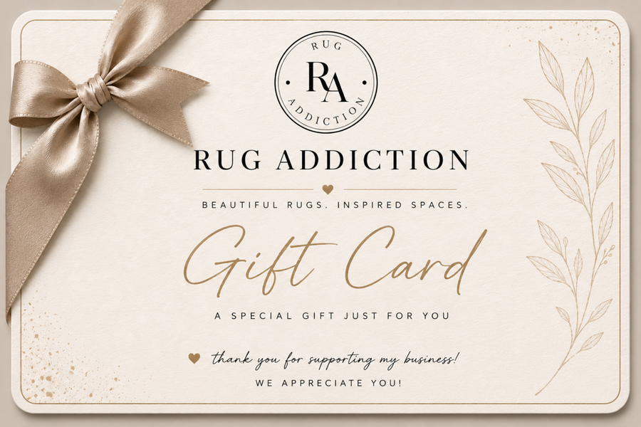 A square gift card from Rug Addiction with a beige satin ribbon bow in the top left corner. The card has a cream background with elegant black and gold typography, featuring the brand logo at the top and decorative botanical line art on the right side.